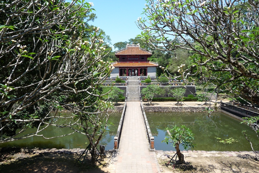 A tiled-roof pavilion at the end of a paved causeway between two ponds, framed by frangipani branches at a Hue royal tomb