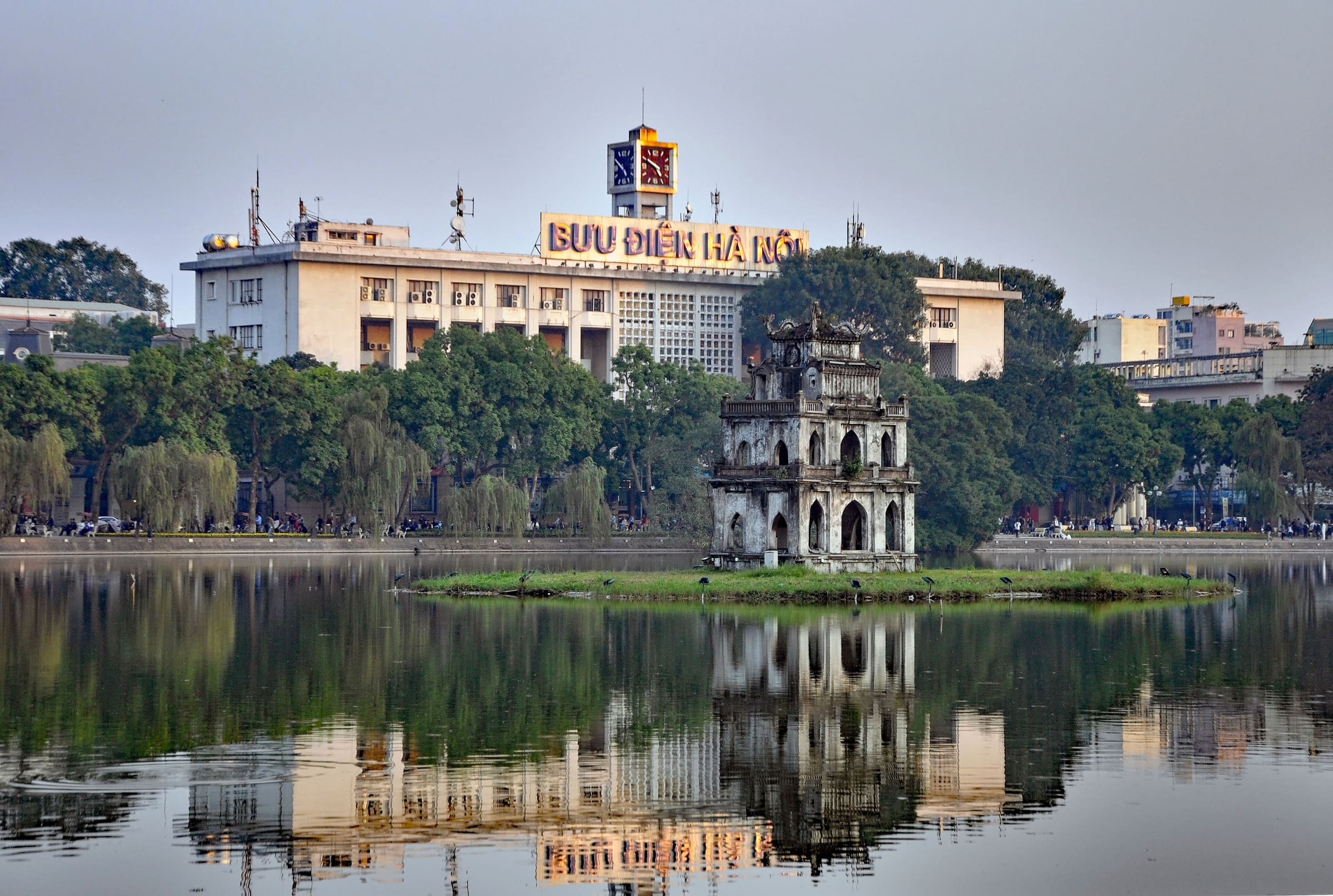 Turtle Tower on its islet in Hoan Kiem Lake, reflected in the still water with the city behind