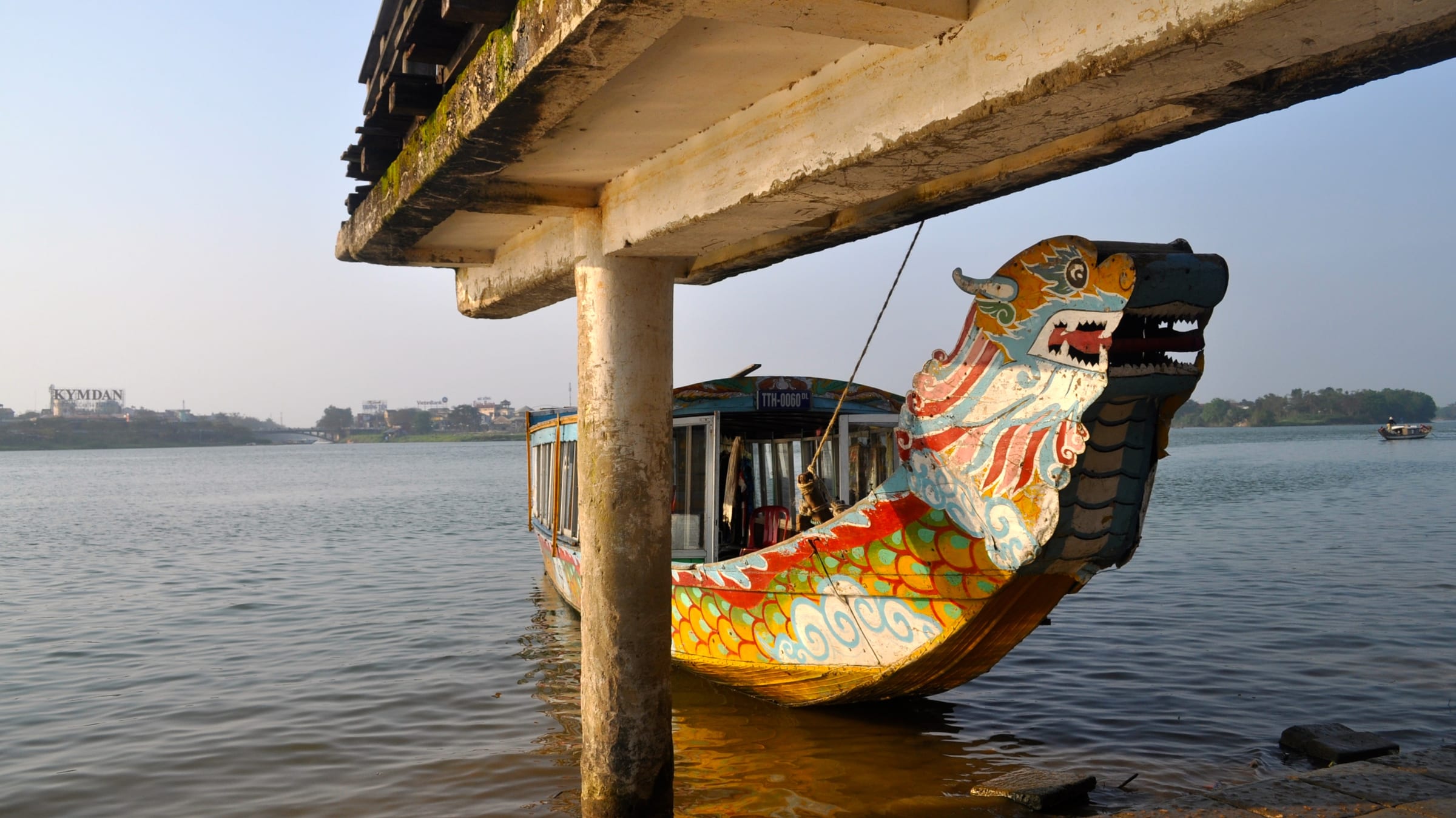 A dragon boat with a golden carved prow moored under a wooden jetty on the Perfume River in Hue